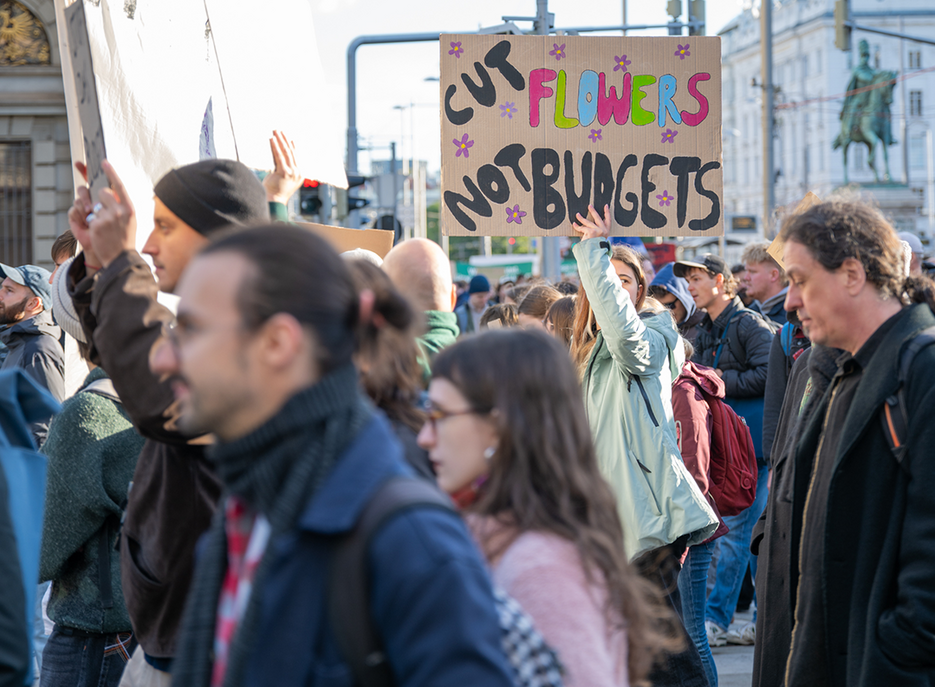 Menschenmenge bei einer Demonstration in der Stadt; eine Person hält ein Schild mit der Aufschrift „Cut Flowers Not Budgets“ über den Kopf. 