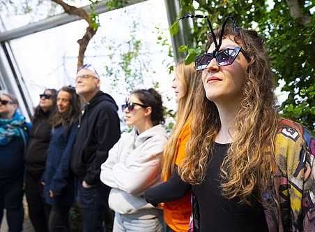 Ein Foto einer Gruppe von Menschen, die in einer Schlange stehen. Eine Person trägt eine schwarze Sonnenbrille und hat lange Haare mit Bienenflügeln auf dem Kopf. Das Foto wurde von der Seite aufgenommen, im Hintergrund ist ein Gewächshaus zu sehen. ©Uni Graz/Nadja Fuchs