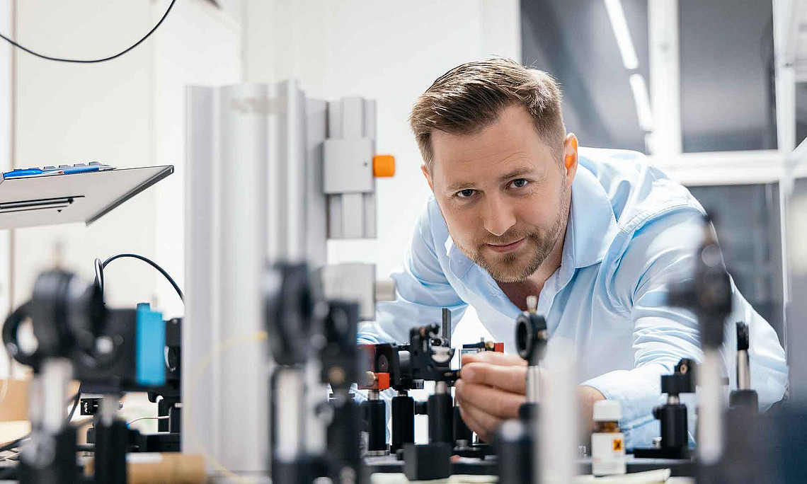 Mann mit kurzem Bart und hellem Hemd arbeitet an optischen Geräten auf einem Labortisch in einem hellen Raum mit Fenster.