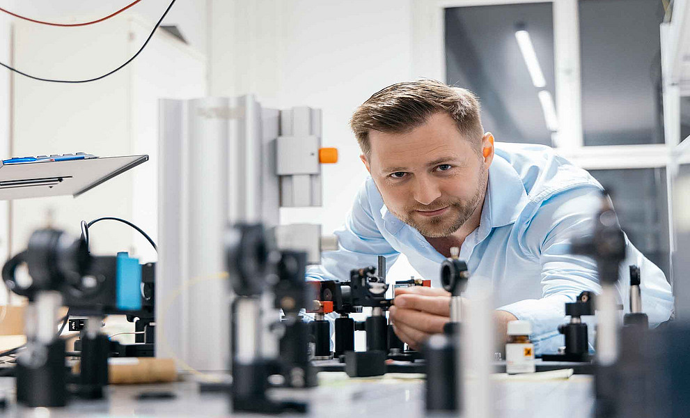 Mann mit kurzem Bart und hellem Hemd arbeitet an optischen Geräten auf einem Labortisch in einem hellen Raum mit Fenster.