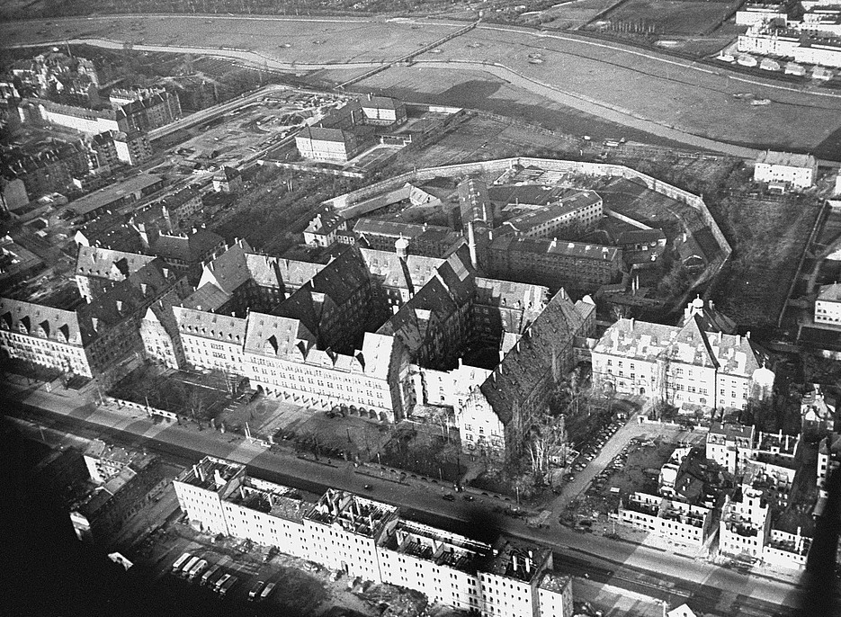 Schwarz-weiß-Luftaufnahme des Justizpalasts in Nürnberg und der angrenzenden Gebäude. Im Vordergrund sind die großen, historischen Gebäudekomplexe mit markanten Dächern zu sehen, dahinter ein achteckiger Gefängnishof, umgeben von einer Mauer. Im Hintergrund erstrecken sich Straßen, Felder und weitere Stadtteile. 