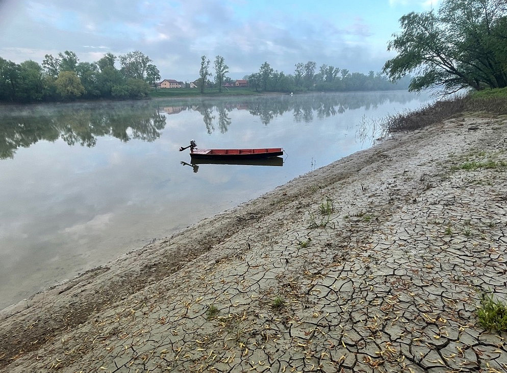 A small red boat floats on a calm river beside a wide, cracked, dry shoreline with trees and houses reflected in the still water. ©@Yvonne Živković
