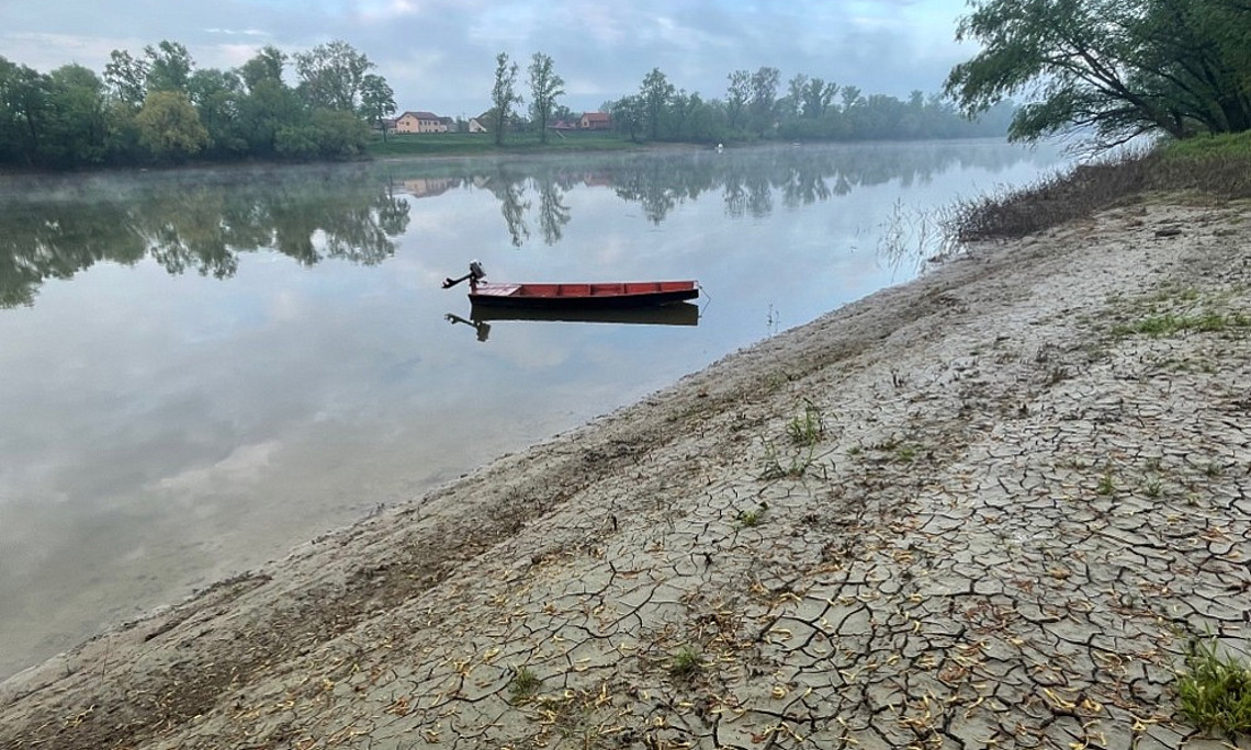 A small red boat floats on a calm river beside a wide, cracked, dry shoreline with trees and houses reflected in the still water.