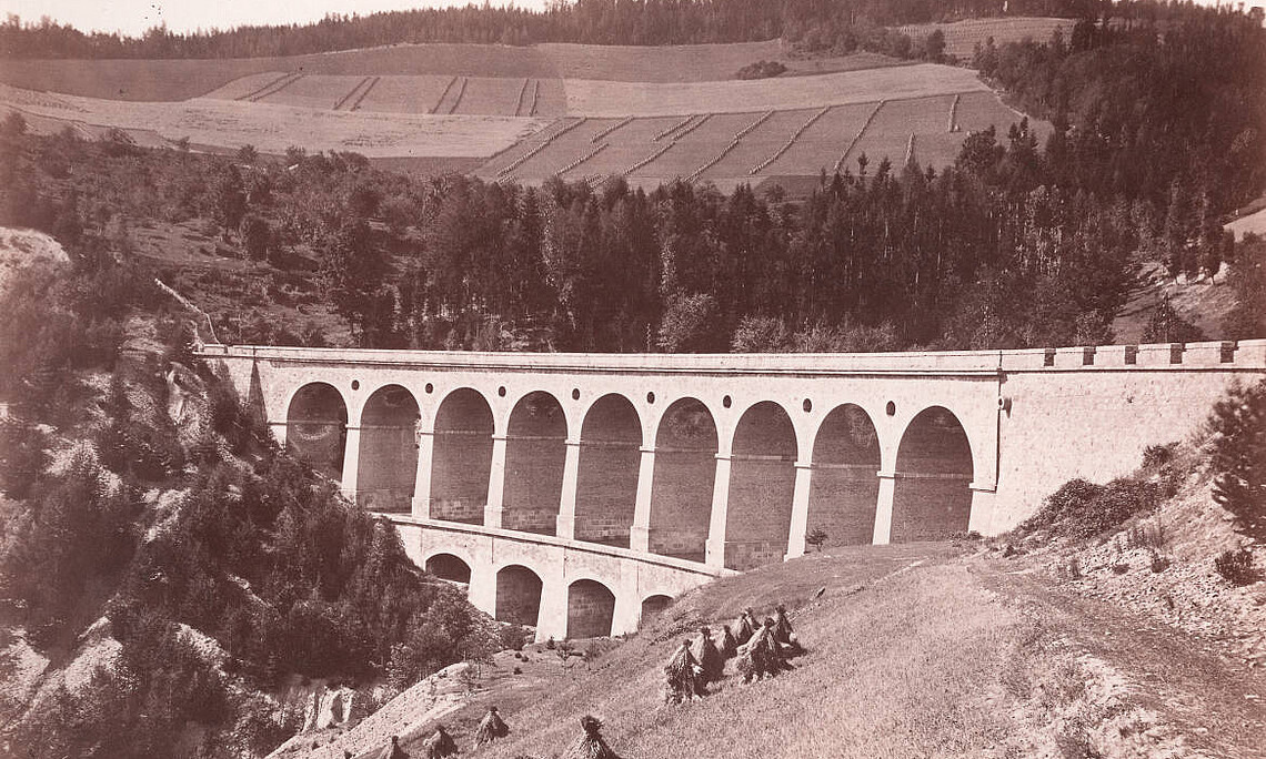 Historic black and white photograph of a viaduct with several large round arches stretching across a wooded ravine. Haystacks in a meadow can be seen in the foreground, fields and a forest on rolling hills in the background