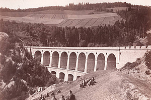 Historic black and white photograph of a viaduct with several large round arches stretching across a wooded ravine. Haystacks in a meadow can be seen in the foreground, fields and a forest on rolling hills in the background