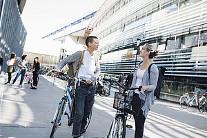 Two young women, each with a bicycle, talking to each other, behind them the Resowi Centre of the University of Graz