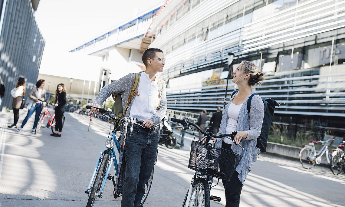 Two young women, each with a bicycle, talking to each other, behind them the Resowi Centre of the University of Graz