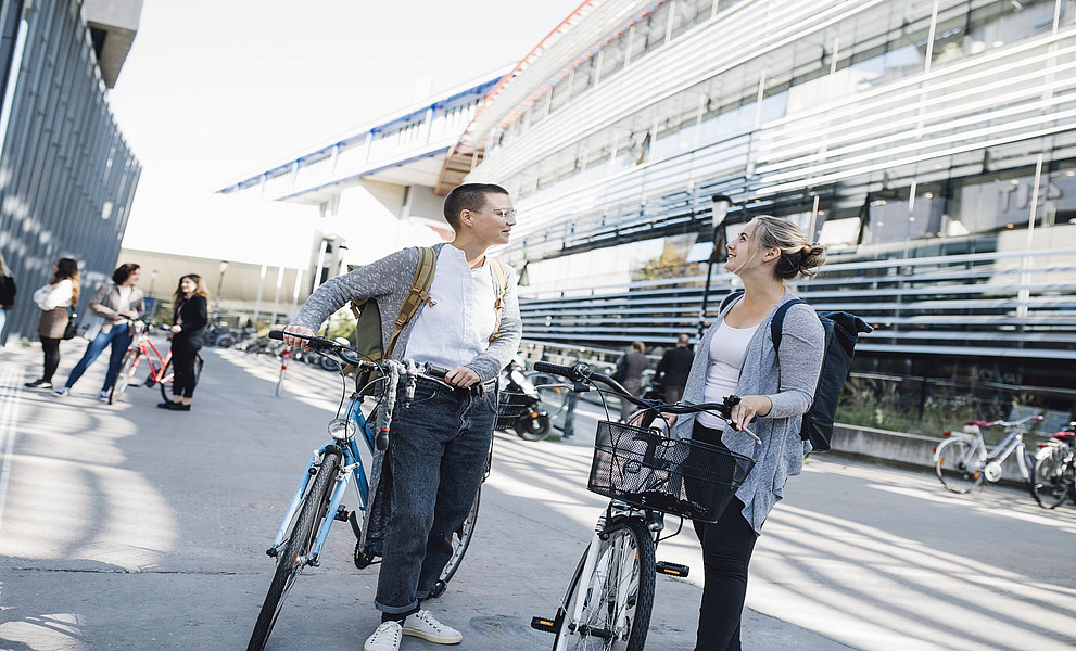 zwei junge Frauen mit jeweils einem Fahrrad sprechen miteinander, dahinter das Resowi-Zentrum der Uni Graz