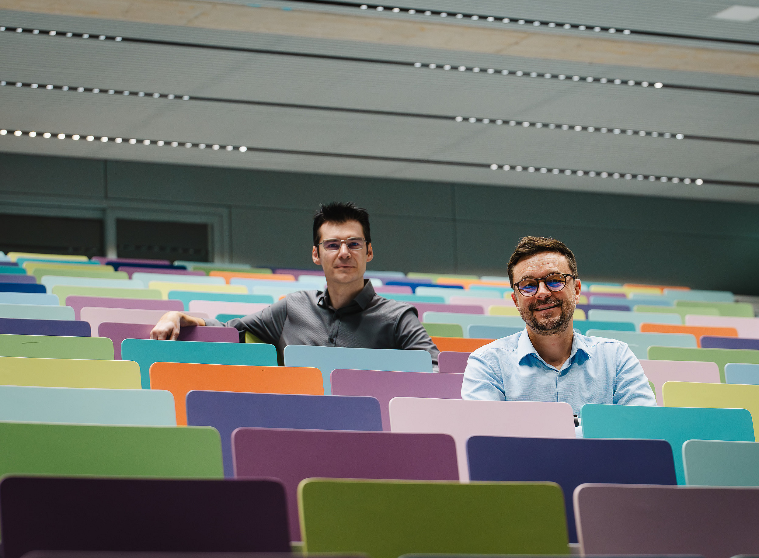 Zwei Männer sitzen inmitten eines leeren Hörsaals einer Universität auf bunten Stühlen und lächeln in die Kamera. ©Uni Graz/Tzivanopoulos