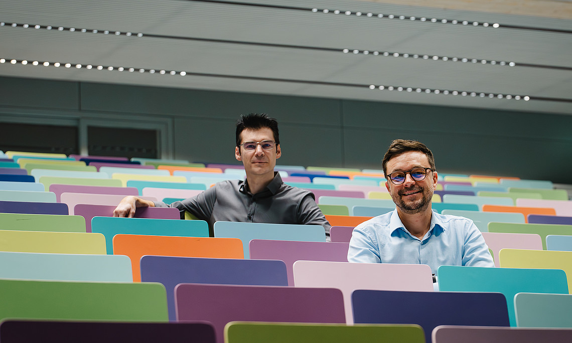Two men sit in the middle of an empty university lecture hall, on colorful chairs and smiling at the camera. 