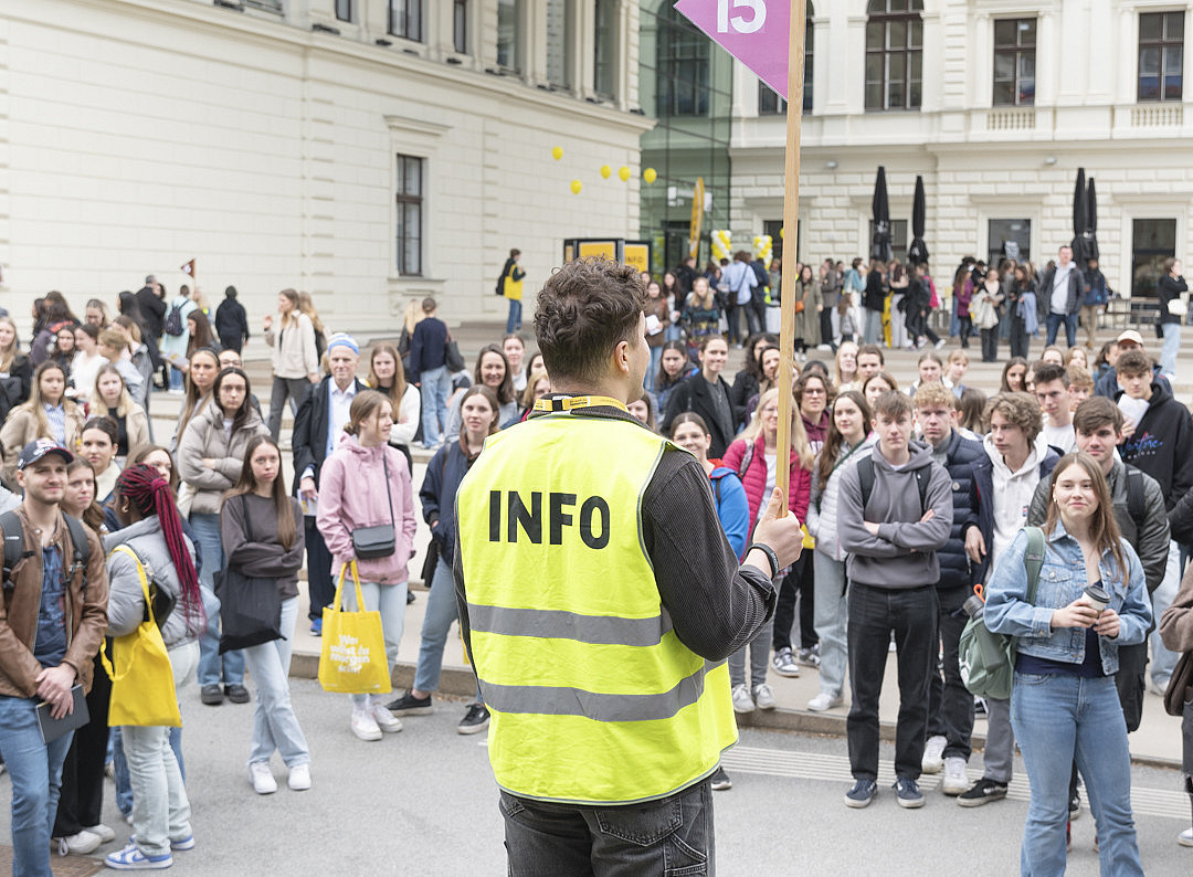 Ein Mann mit einer Veste, auf der "Info" steht und eine Gruppe von jungen Menschen, die um ihn stehen. ©Uni Graz/Angele