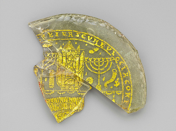 Dieses seltene Beispiel jüdischen Goldglases zeigt eine offene Thora-Arche mit gerollten Schriftrollen auf ihren Regalen und rituellen Geräten des Tempels - darunter zwei Menorot (Leuchter), ein Schofar (Widderhorn) und ein Etrog (Zitrone). Ursprünglich war darunter eine Festmahlszene mit einem Fisch auf dem Dreifußtisch vor einem Kissen dargestellt. ©The Metropolitan Museum of Art. "Bowl Fragments with Menorah, Shofar, and Torah Ark" Inv. 18.145.1a, b