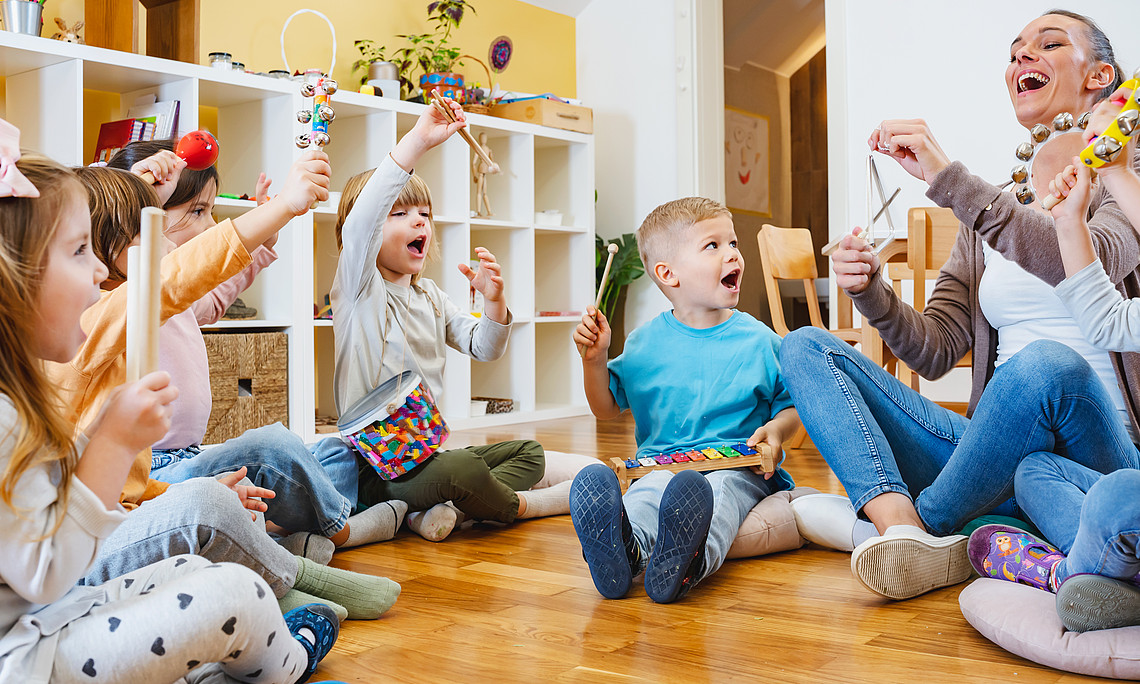 Eine Gruppe von Kindern spielt im Kindergarten mit Musikinstrumenten, während ihre Lehrerin auf dem Boden sitzt und mitsingt. Der Raum hat weiße Wände und Holzböden und ist mit bunten Spielsachen wie Küchenutensilien und Spielgeräten gefüllt.
