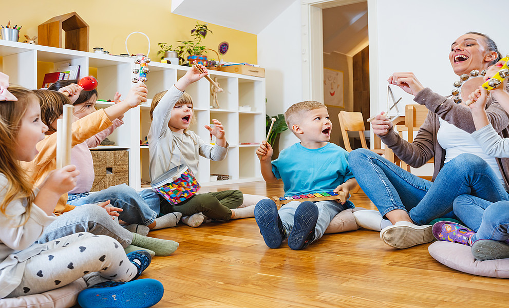 Eine Gruppe von Kindern spielt im Kindergarten mit Musikinstrumenten, während ihre Lehrerin auf dem Boden sitzt und mitsingt. Der Raum hat weiße Wände und Holzböden und ist mit bunten Spielsachen wie Küchenutensilien und Spielgeräten gefüllt.