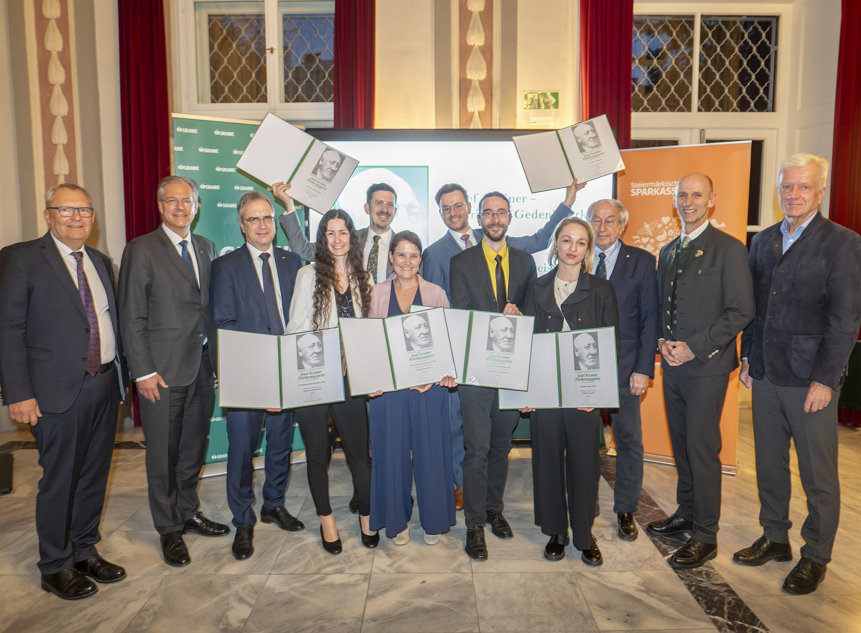 Eine Gruppe von Personen steht in einem festlichen Innenraum mit hellem Marmorboden und hohen Fenstern, die von roten Vorhängen eingerahmt sind. Alle tragen formelle Kleidung wie Anzüge, Kostüme oder elegante Hosen und Blusen. Mehrere Personen in der Mitte halten große, aufgeklappte Urkundenmappen in die Kamera, einige heben sie über den Kopf. Im Hintergrund stehen zwei Roll‑up‑Banner mit Logos und Schriftzügen sowie ein Bildschirm, der auf eine Preis‑ oder Ehrungsveranstaltung schließen lässt. ©Foto Fischer