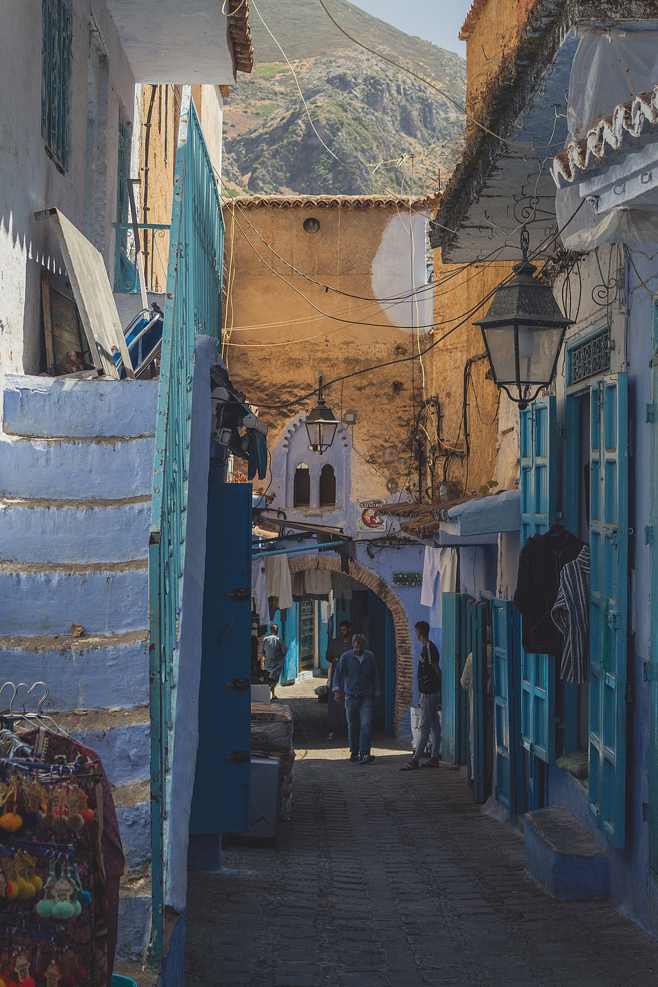 Eine kleine Gasse mit blau, türkis und weiß gestrichenen Häusern in Chefchaouen, Marokko; im Hintergrund sind Berge zu sehen.