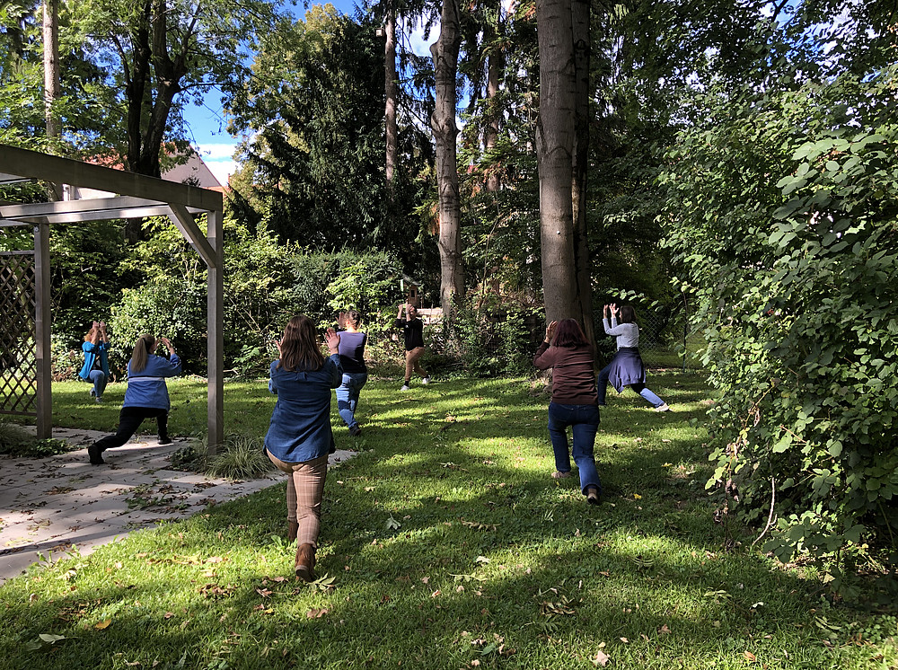 Mehrere Personen machen Übungen in einem Garten. Sie stehen im Ausfallschritt auf dem Rasen. Im Hintergrund sind Bäume und Sträucher. Auf der linken Seite sieht man Teile einer Laube. 