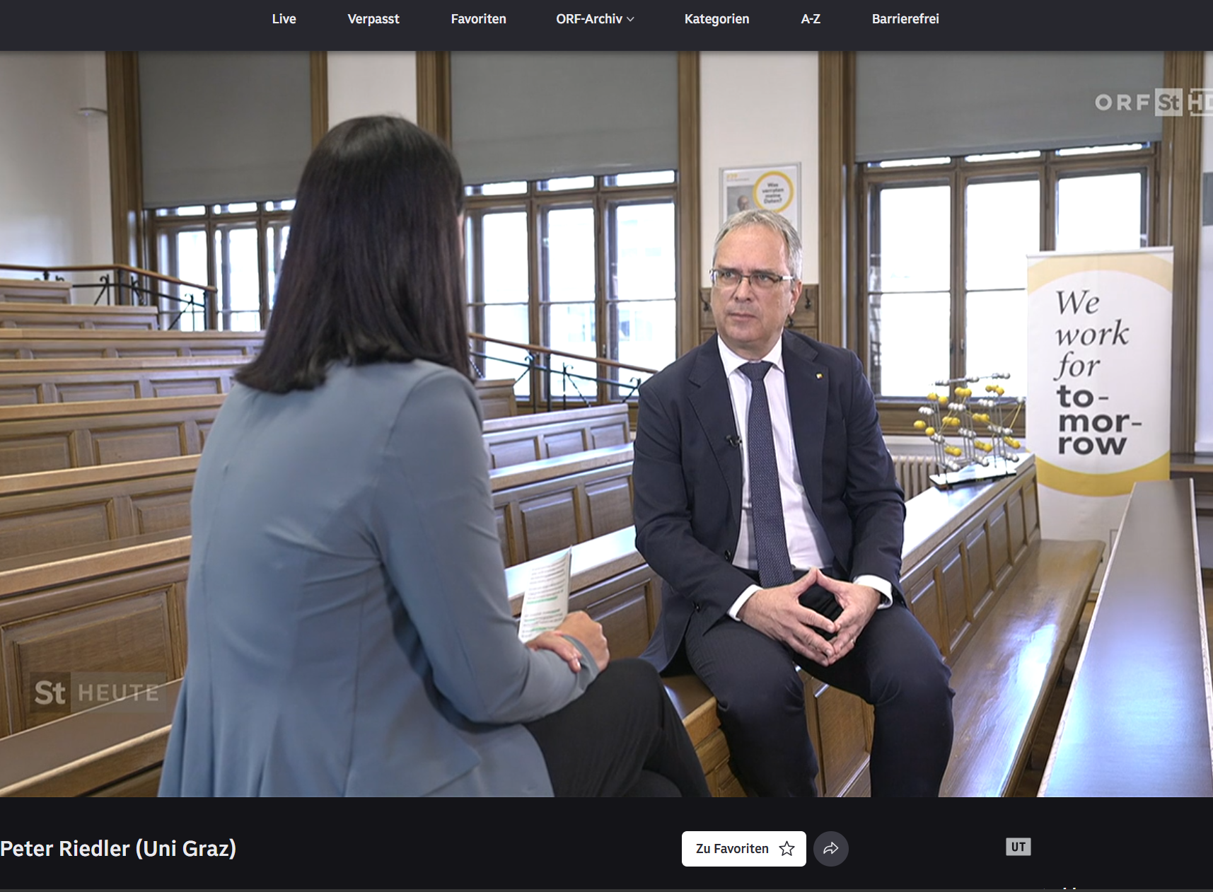 Mann mittleren Alters im dunklen Anzug sitzt in einem Hörsaal und spricht mit Frau in hellblauem Blazer, Banner „We work for tomorrow“ im Hintergrund 
