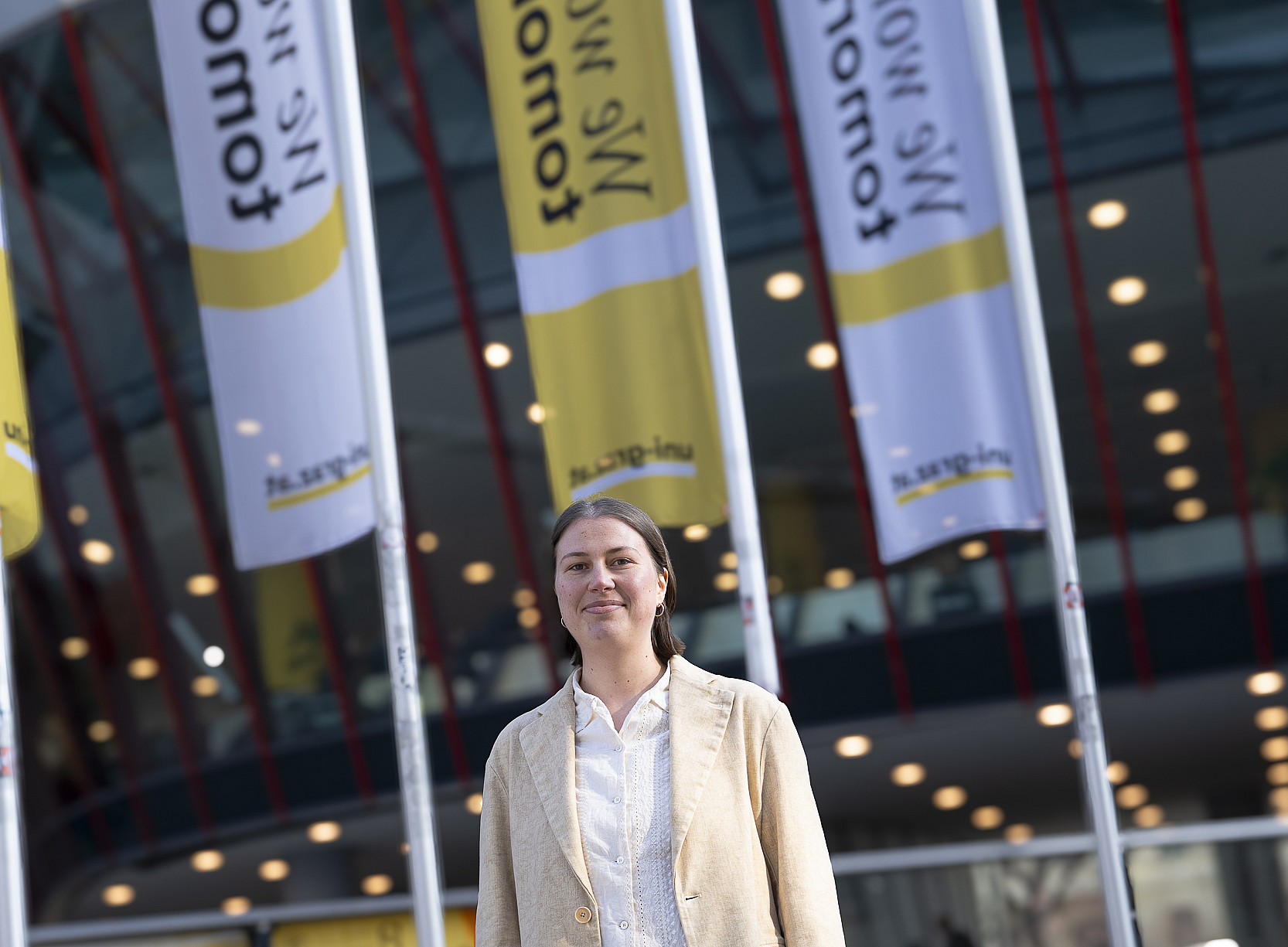 A person wearing a light beige blazer and white blouse stands outdoors in front of a modern glass building, with several towering yellow and white flags bearing inscriptions in the background and many round ceiling lights in the foyer of the building. ©Uni Graz/Tzivanopoulos