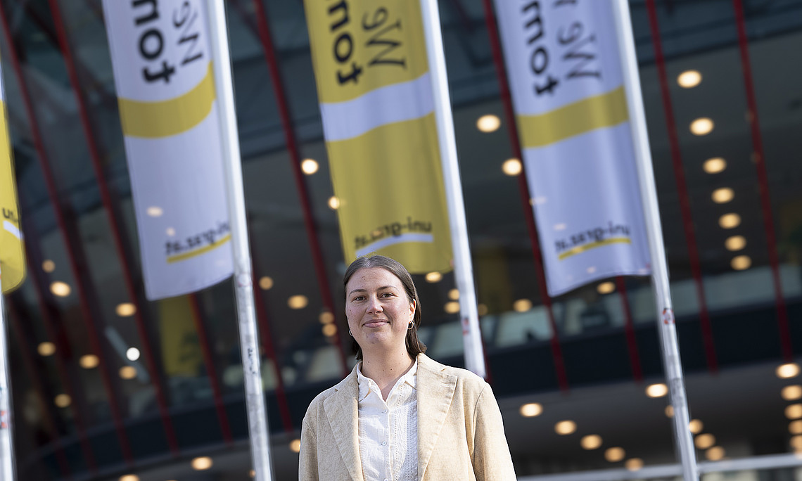 A person wearing a light beige blazer and white blouse stands outdoors in front of a modern glass building, with several towering yellow and white flags bearing inscriptions in the background and many round ceiling lights in the foyer of the building.