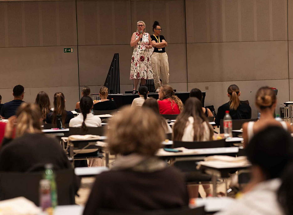 Zwei Frauen mittleren Alters stehen auf einer Bühne, sprechen vor sitzendem Publikum aus jungen Erwachsenen in einer Halle. ©Uni Graz/Angele