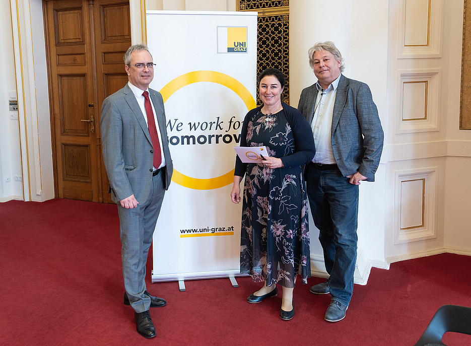 Drei Personen stehen in einem repräsentativen Innenraum mit rotem Teppich vor einem Roll‑Up‑Banner der Uni Graz. Auf dem Banner ist ein gelber Kreis und der Slogan „We work for tomorrow“ sowie die Webadresse www.uni-graz.at zu sehen. Links steht eine Person im grauen Anzug mit roter Krawatte, in der Mitte eine Person im dunklen, geblümten Kleid, die ein Folder oder Heft in der Hand hält, rechts eine Person in Jeans, Hemd und grauem Sakko. Im Hintergrund sind eine Holztür und verzierte Wandflächen zu erkennen. ©Uni Graz/Angele