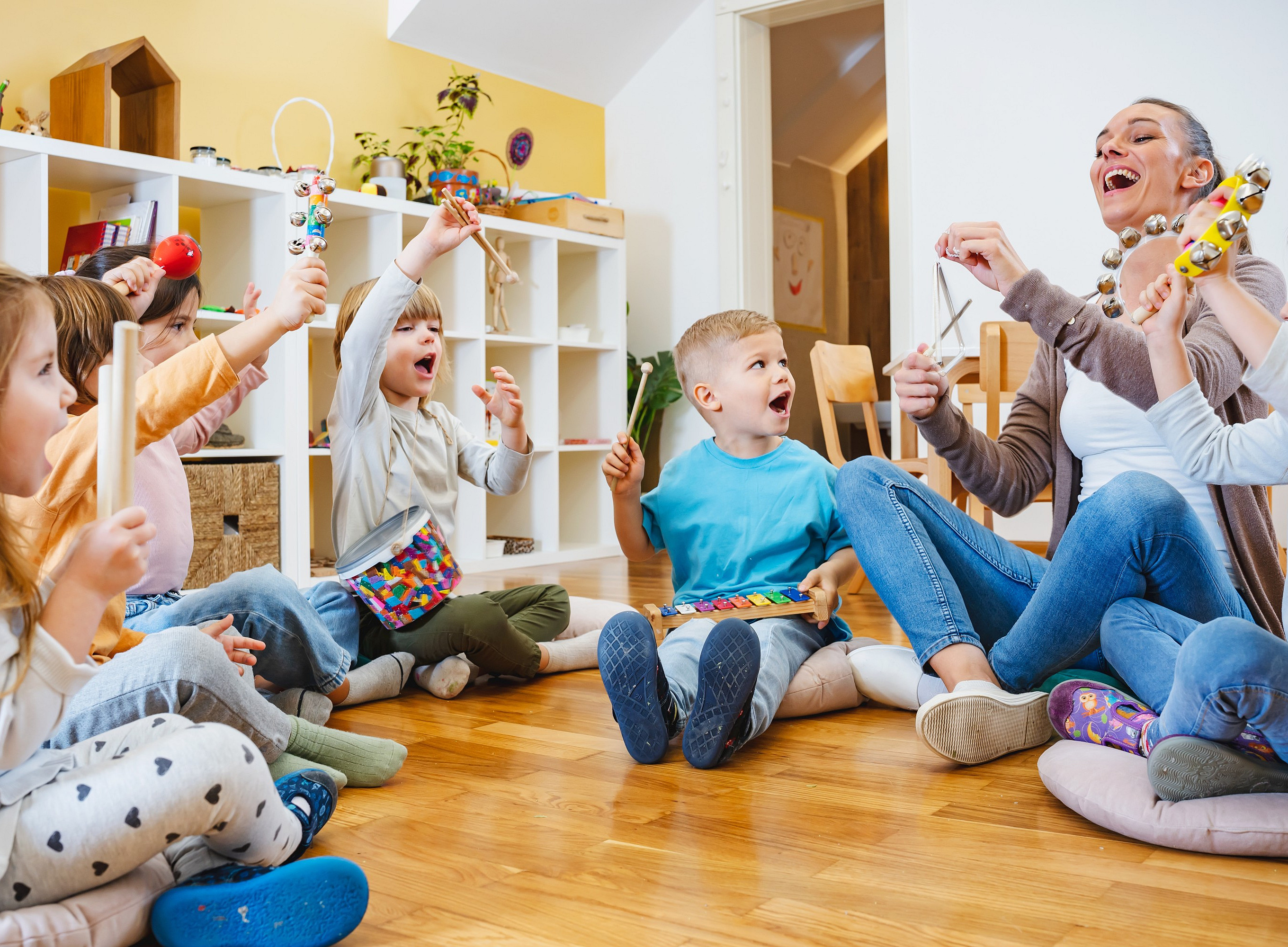 Mehrere Kindergartenkinder und eine erwachsene Person sitzen im Kreis auf Kissen und spielen gemeinsam verschiedene Musikinstrumente im Gruppenraum. 