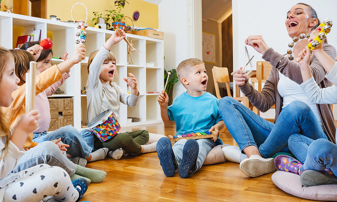 Mehrere Kindergartenkinder und eine erwachsene Person sitzen im Kreis auf Kissen und spielen gemeinsam verschiedene Musikinstrumente im Gruppenraum.