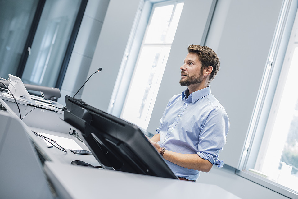 Professor steht im Hörsaal hinter dem Pult und spricht; sein Blick ist Richtung Mitte des Raumes gerichtet