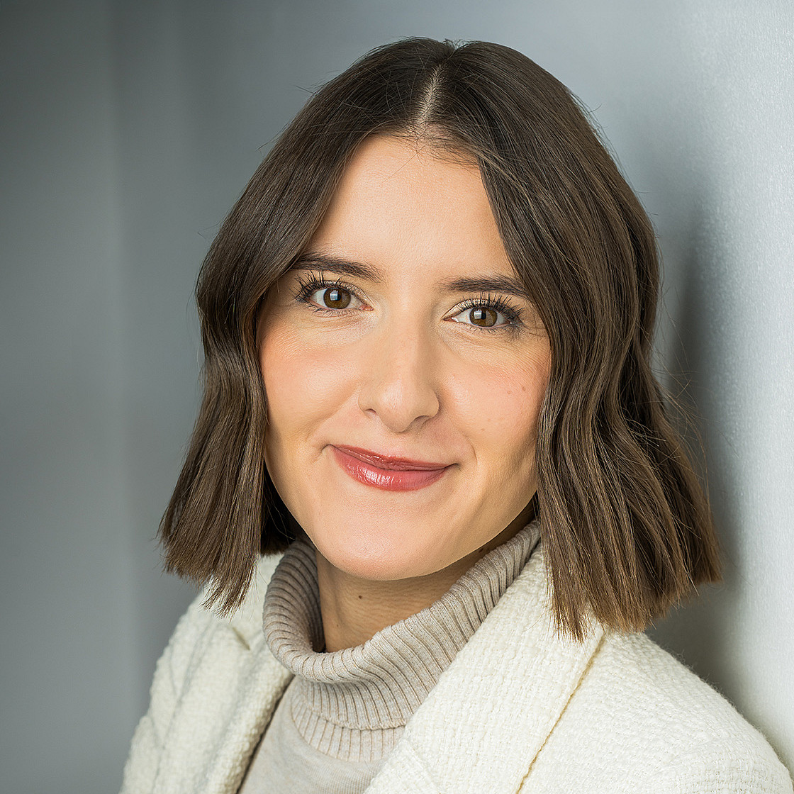 Porträt einer jungen Frau mit schulterlangen brauchen Haaren und braunen Augen, die einen weißen Blazer trägt und mit verschränkten Armen in die Kamera schaut