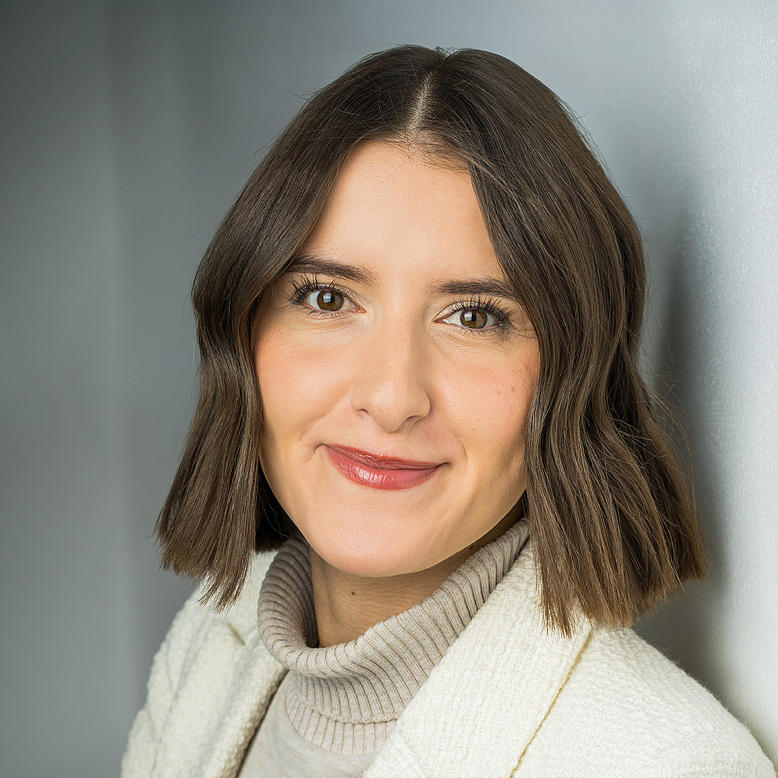 Porträt einer jungen Frau mit schulterlangen brauchen Haaren und braunen Augen, die einen weißen Blazer trägt und mit verschränkten Armen in die Kamera schaut