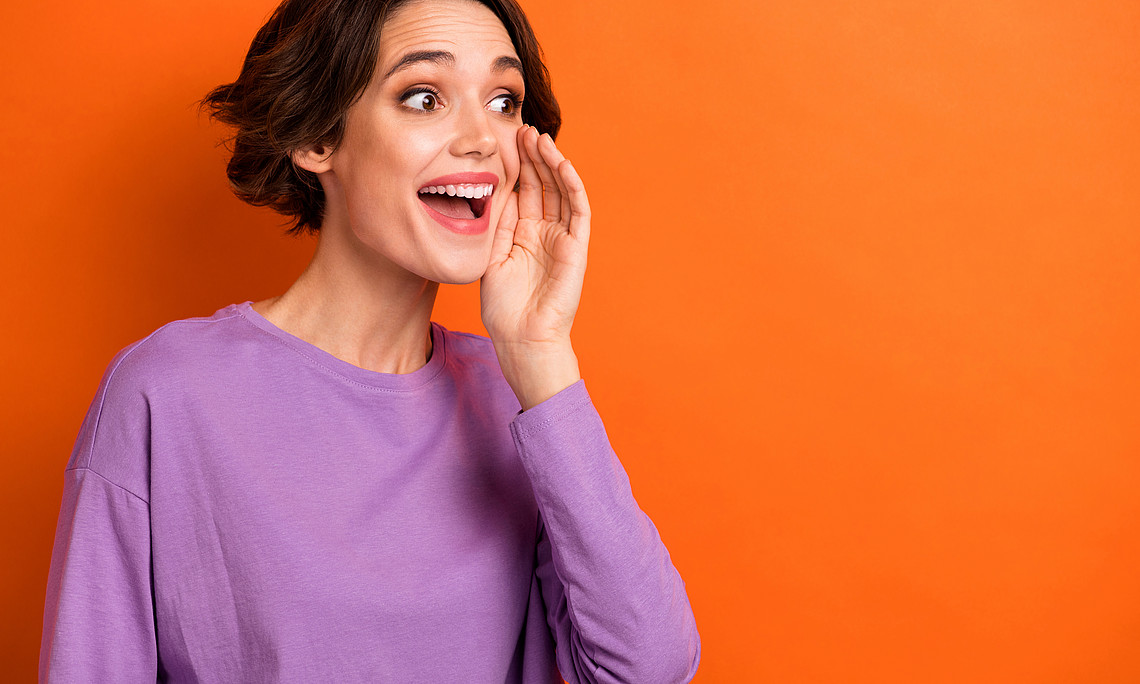 Junge Frau mit sehr erfreutem Gesicht vor orangen Hintergrund formt mit einer Hand einen Lautsprecher und ruft eine Ankündigung aus