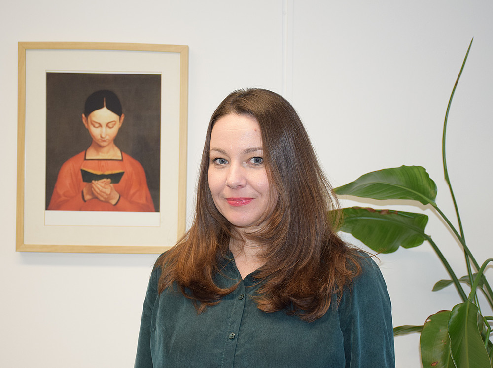 Portrait of historian Tanja Skambraks in her office. In the background, you can see the painting "Lesendes Mädchen" (Girl Reading) by ustav Adolph Hennig and a green plant. ©Ulrike Freitag, Universität Graz