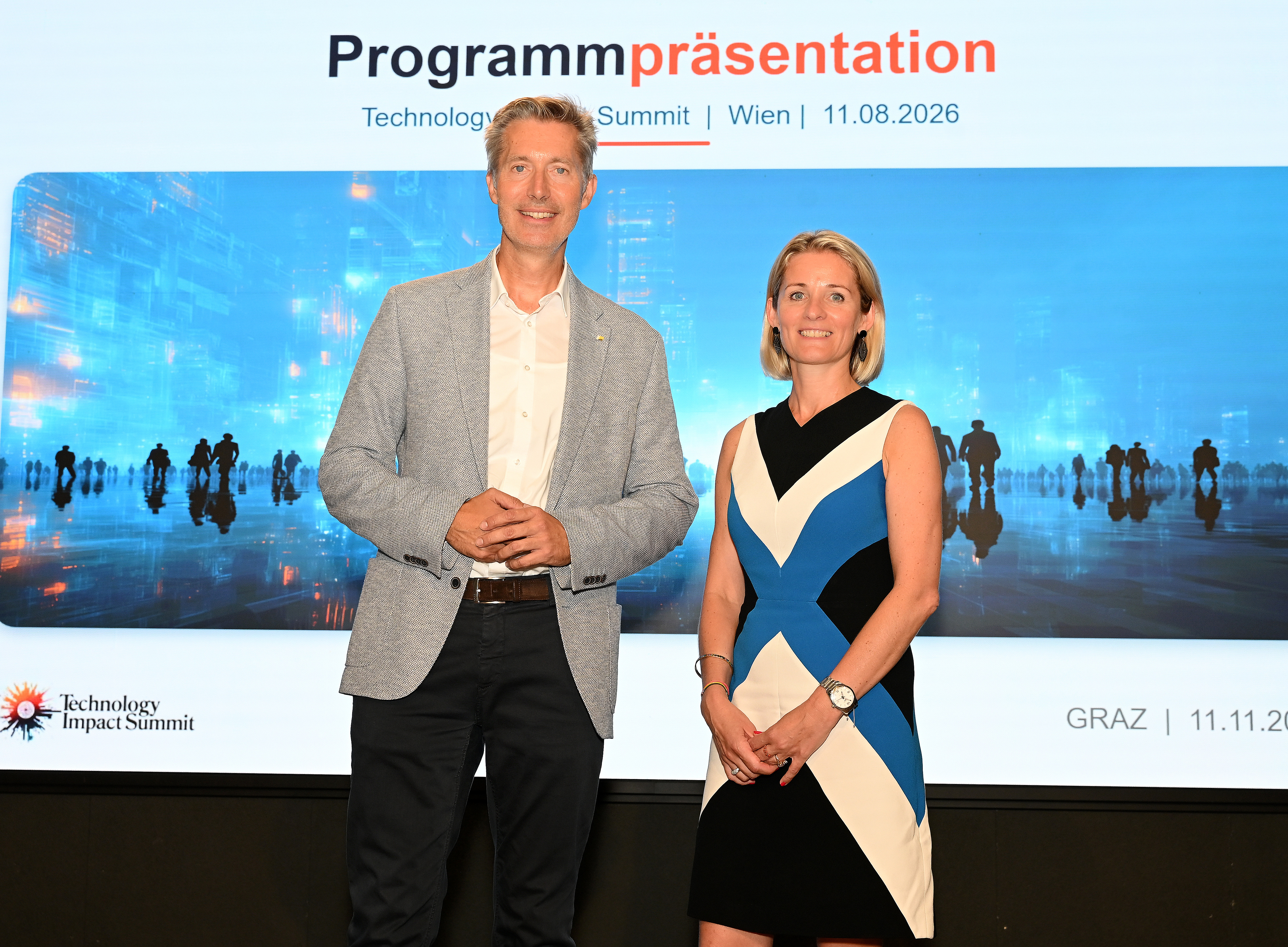 Mann im grauen Blazer und Frau im schwarz-blau-weißen Kleid stehen lächelnd vor Leinwand mit Aufschrift „Programmpräsentation Technology Impact Summit“. 