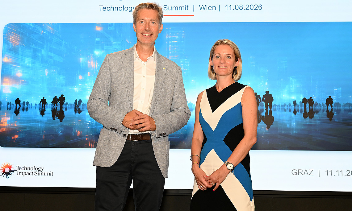 Mann im grauen Blazer und Frau im schwarz-blau-weißen Kleid stehen lächelnd vor Leinwand mit Aufschrift „Programmpräsentation Technology Impact Summit“.