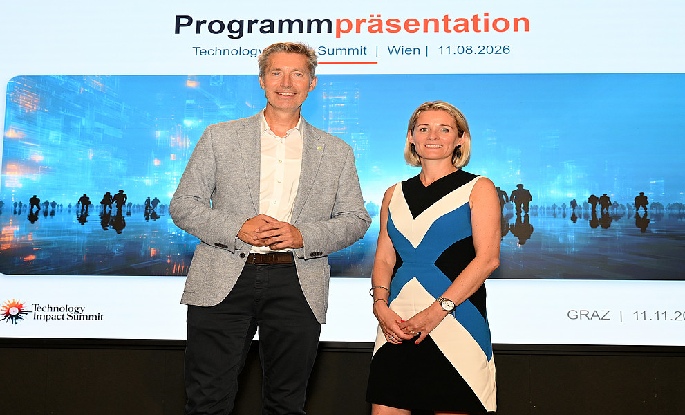 Mann im grauen Blazer und Frau im schwarz-blau-weißen Kleid stehen lächelnd vor Leinwand mit Aufschrift „Programmpräsentation Technology Impact Summit“.