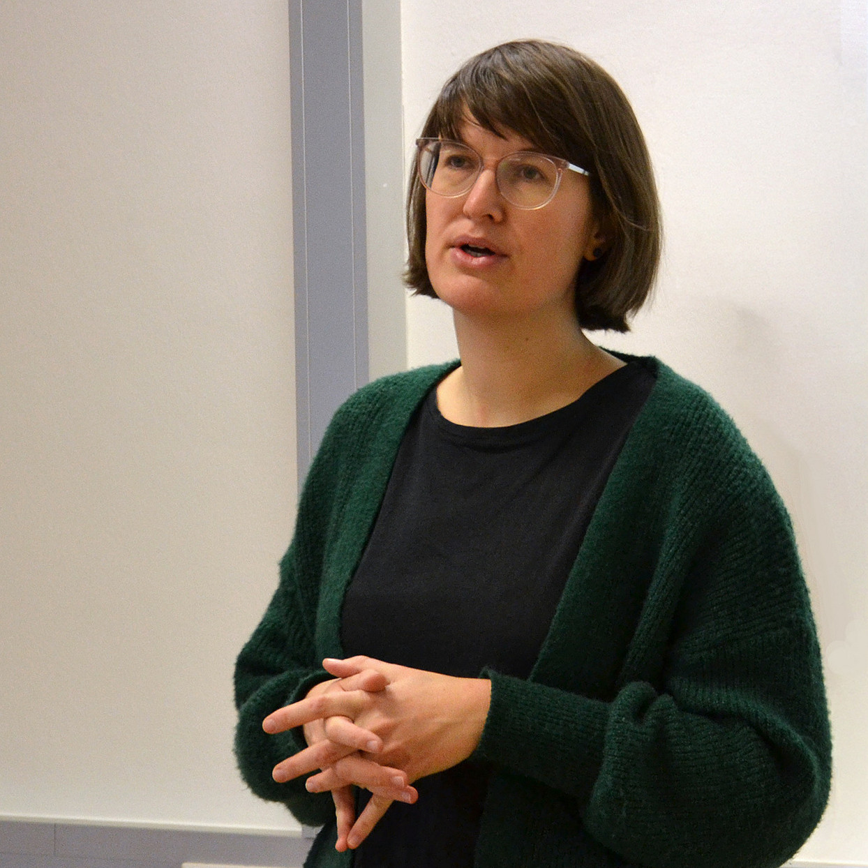 eine Frau mit schulterlangen braunen Haaren und Brille, die vor einer weißen Wand steht und spricht