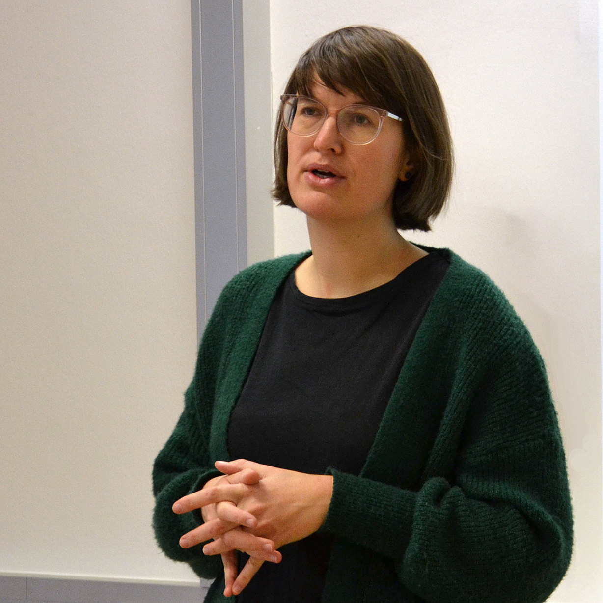 eine Frau mit schulterlangen braunen Haaren und Brille, die vor einer weißen Wand steht und spricht