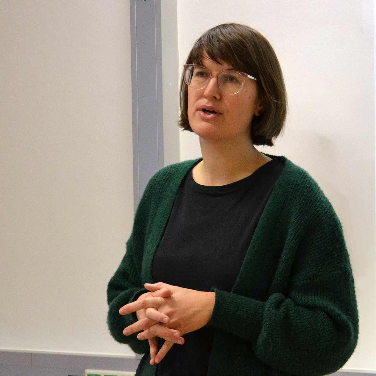 eine Frau mit schulterlangen braunen Haaren und Brille, die vor einer weißen Wand steht und spricht