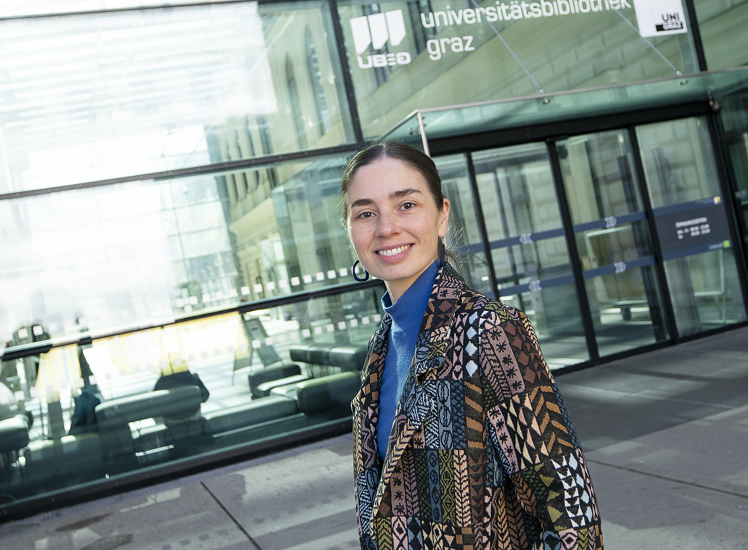 Person in semi-close-up profile standing in front of a modern glass facade with a large entrance area; wearing a patterned patchwork coat in earth tones over a blue turtleneck jumper, one hand in the coat pocket; reflections of the interior and exterior on the glass panes, clear, urban background. ©Uni Graz/Tzivanopoulos