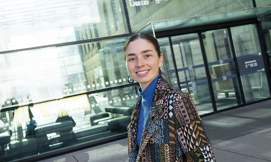 Person in semi-close-up profile standing in front of a modern glass facade with a large entrance area; wearing a patterned patchwork coat in earth tones over a blue turtleneck jumper, one hand in the coat pocket; reflections of the interior and exterior on the glass panes, clear, urban background.