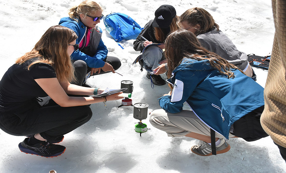 Eine Gruppe junger Frauen und Männer in Outdoor-Kleidung mit blauen Akzenten kniet im Schnee um einen Kessel herum, der zwischen ihnen steht. Jeder hält sein eigenes Notizbuch oder seinen Notizblock in der Hand und macht sich Notizen, während sie zwei Personen dabei zusehen, wie sie mit Eis aus den schmelzenden Gletschern Wasser kochen.