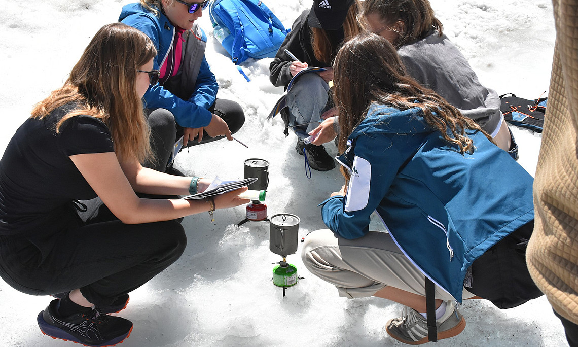A group of young women and men, dressed in outdoor with blue accents, are kneeling on the snow around an kettle placed between them. Each person is holding their own notebook or notepad, taking notes as they watch two people boiling water using ice from the melting glaciers. 