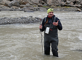 Wolfgang Schöner stands in waders in the drainage channel of a glacier in Greenland, holding a measuring rod in his hand, the device for it hanging around his neck, looking into the camera and speaking.
