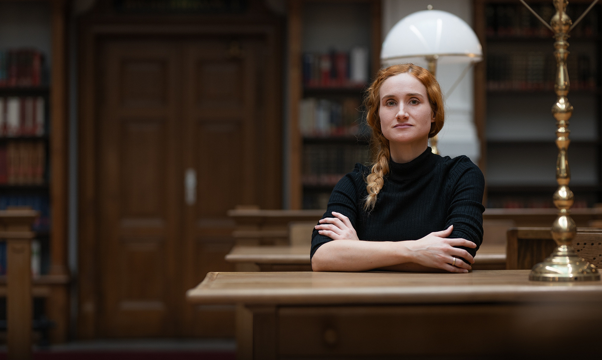 Portrait of a woman with braided hair and a black top, sitting with her arms crossed at a wooden table in a historic reading room. In the background are bookshelves, wooden furniture, and a large brass lamp. ©Uni Graz/Angele