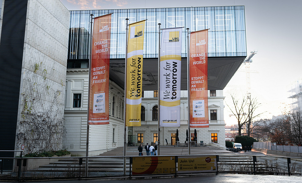 Außenansicht eines modernen Universitätsgebäudes mit Glasfassade und historischem Gebäudeteil. Vor dem Eingang wehen vier große Fahnen: Zwei orangefarbene mit der Aufschrift ‚Orange the World – Stoppt Gewalt an Frauen!‘ und zwei gelb-weiße mit der Aufschrift ‚We work for tomorrow‘. Im Vordergrund ein Geländer mit einem Banner, im Hintergrund ein heller Himmel und Baukräne.
