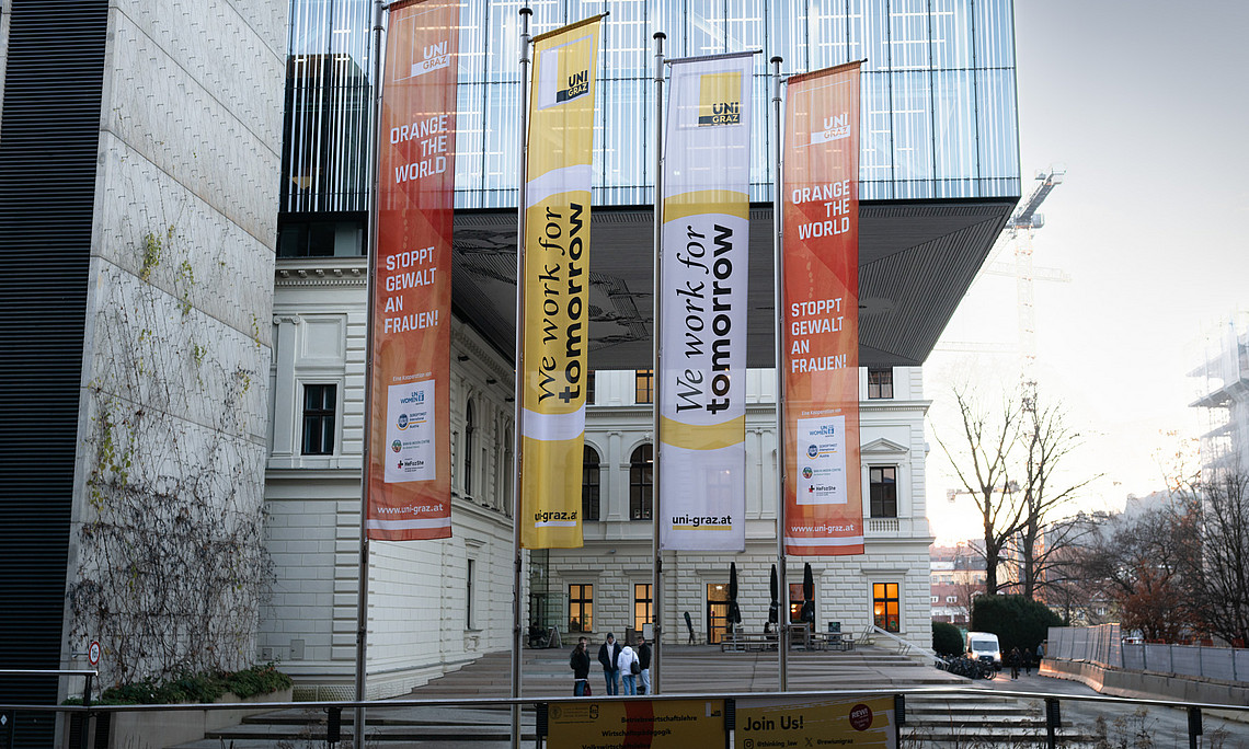 Außenansicht eines modernen Universitätsgebäudes mit Glasfassade und historischem Gebäudeteil. Vor dem Eingang wehen vier große Fahnen: Zwei orangefarbene mit der Aufschrift ‚Orange the World – Stoppt Gewalt an Frauen!‘ und zwei gelb-weiße mit der Aufschrift ‚We work for tomorrow‘. Im Vordergrund ein Geländer mit einem Banner, im Hintergrund ein heller Himmel und Baukräne.