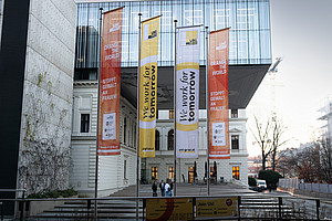 Außenansicht eines modernen Universitätsgebäudes mit Glasfassade und historischem Gebäudeteil. Vor dem Eingang wehen vier große Fahnen: Zwei orangefarbene mit der Aufschrift ‚Orange the World – Stoppt Gewalt an Frauen!‘ und zwei gelb-weiße mit der Aufschrift ‚We work for tomorrow‘. Im Vordergrund ein Geländer mit einem Banner, im Hintergrund ein heller Himmel und Baukräne.