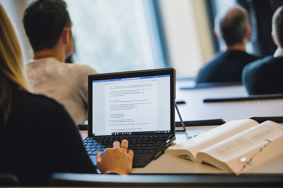 Blick von hinten seitlich auf eine Studentin gerichtet, die im Seminarraum sitzt und auf ihrem Computer rechtliche Inhalte mitschreibt.