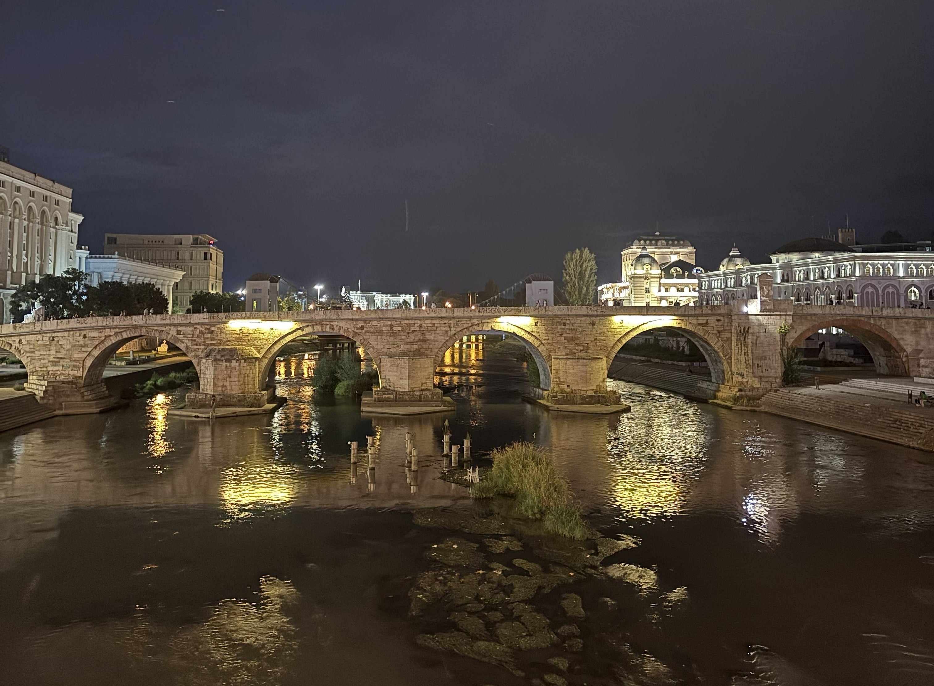 a beautiful night scene of a stone bridge crossing a river. The bridge is illuminated with warm yellow lights, which reflect on the calm water below. The sky is dark, and some buildings on both sides of the river are also lit up, adding to the picturesque atmosphere. ©@ privat Ivana Spirovska